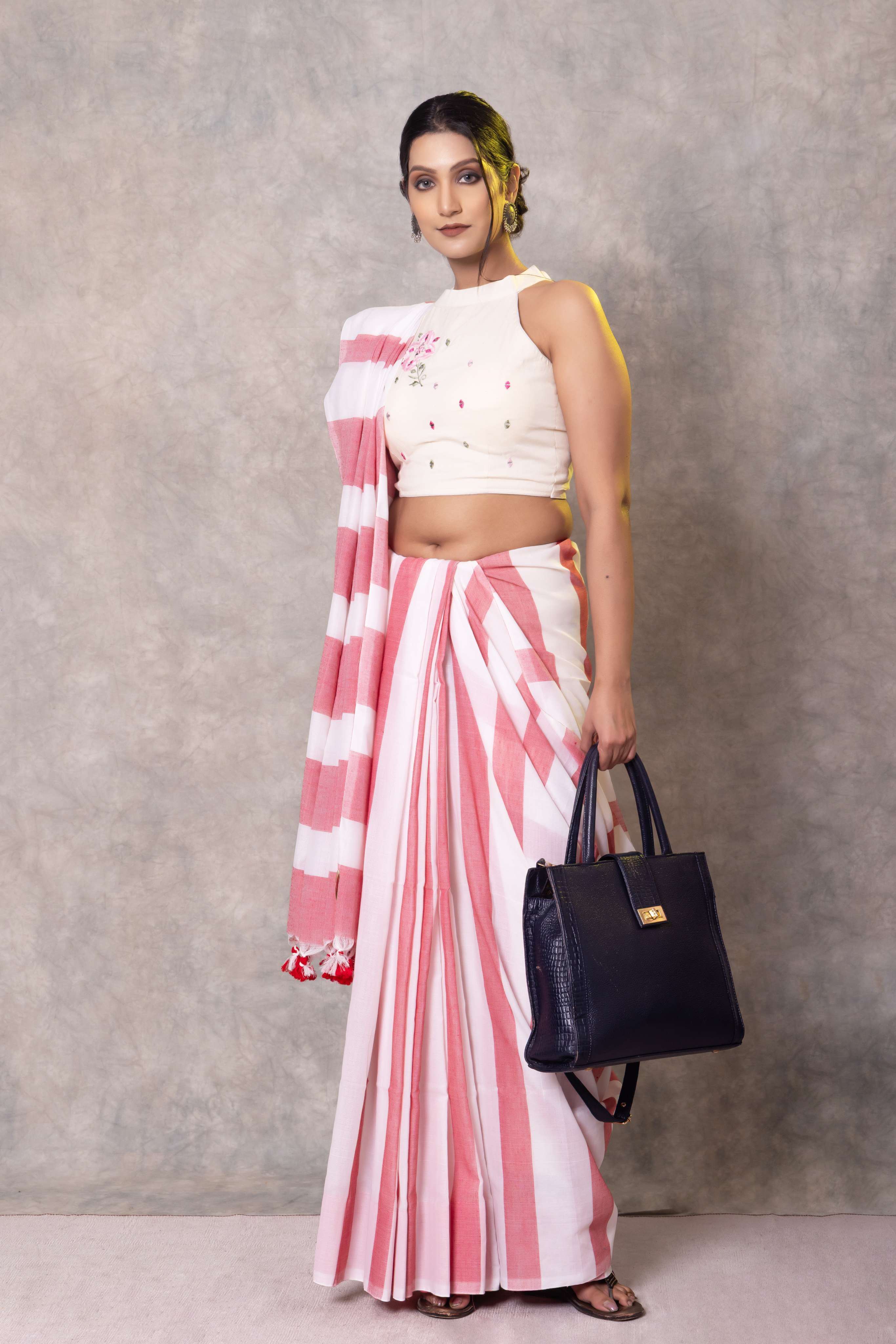 Woman wearing a white and pink striped saree with a black handbag against a gray background