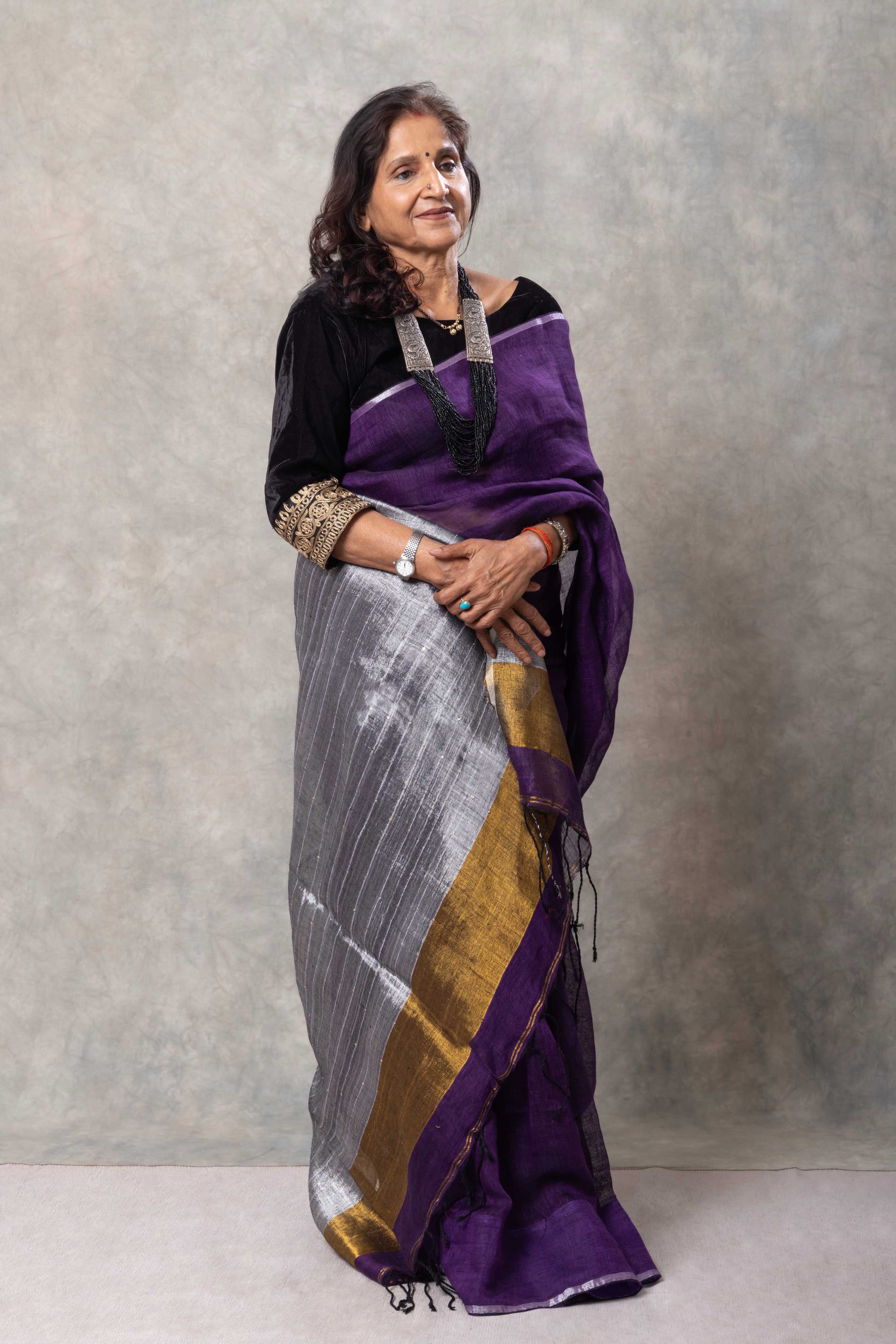 Woman wearing a purple and gray saree with a gold border against a plain background
