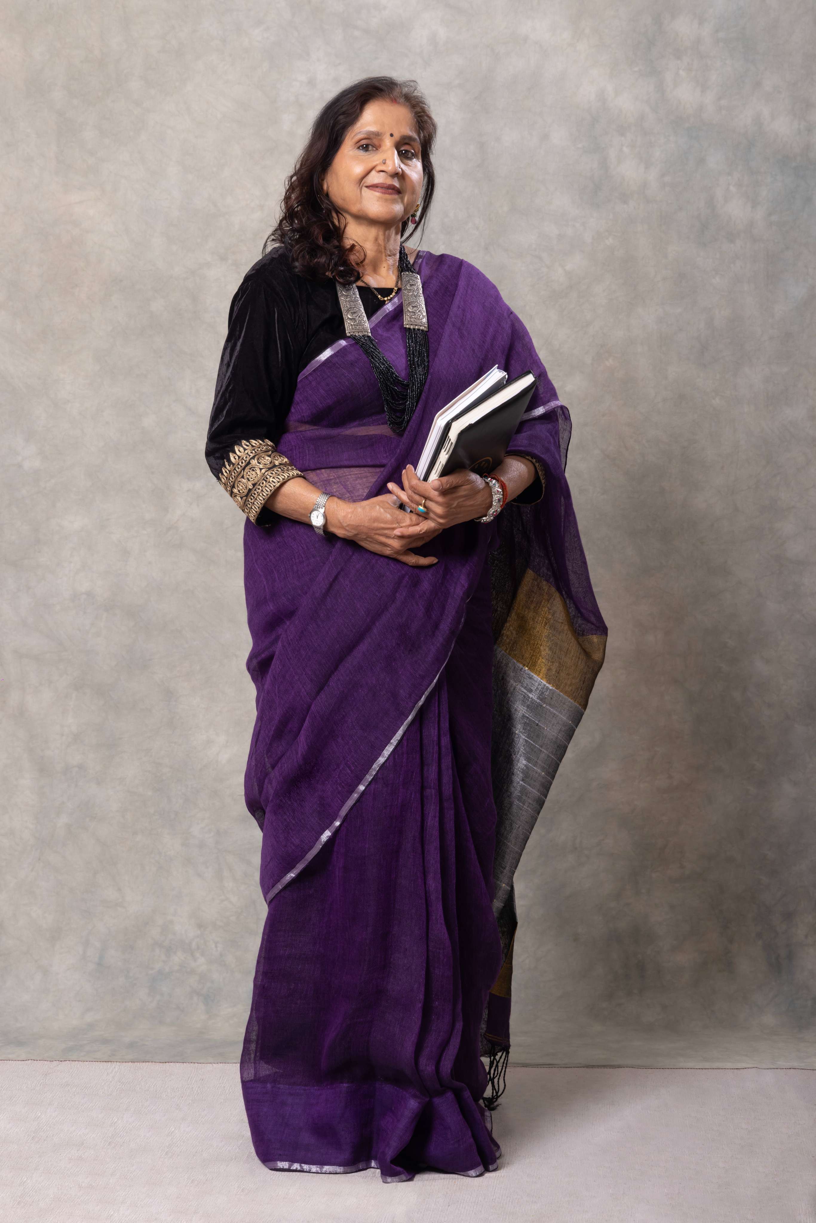 Woman in a purple saree holding a book against a plain background