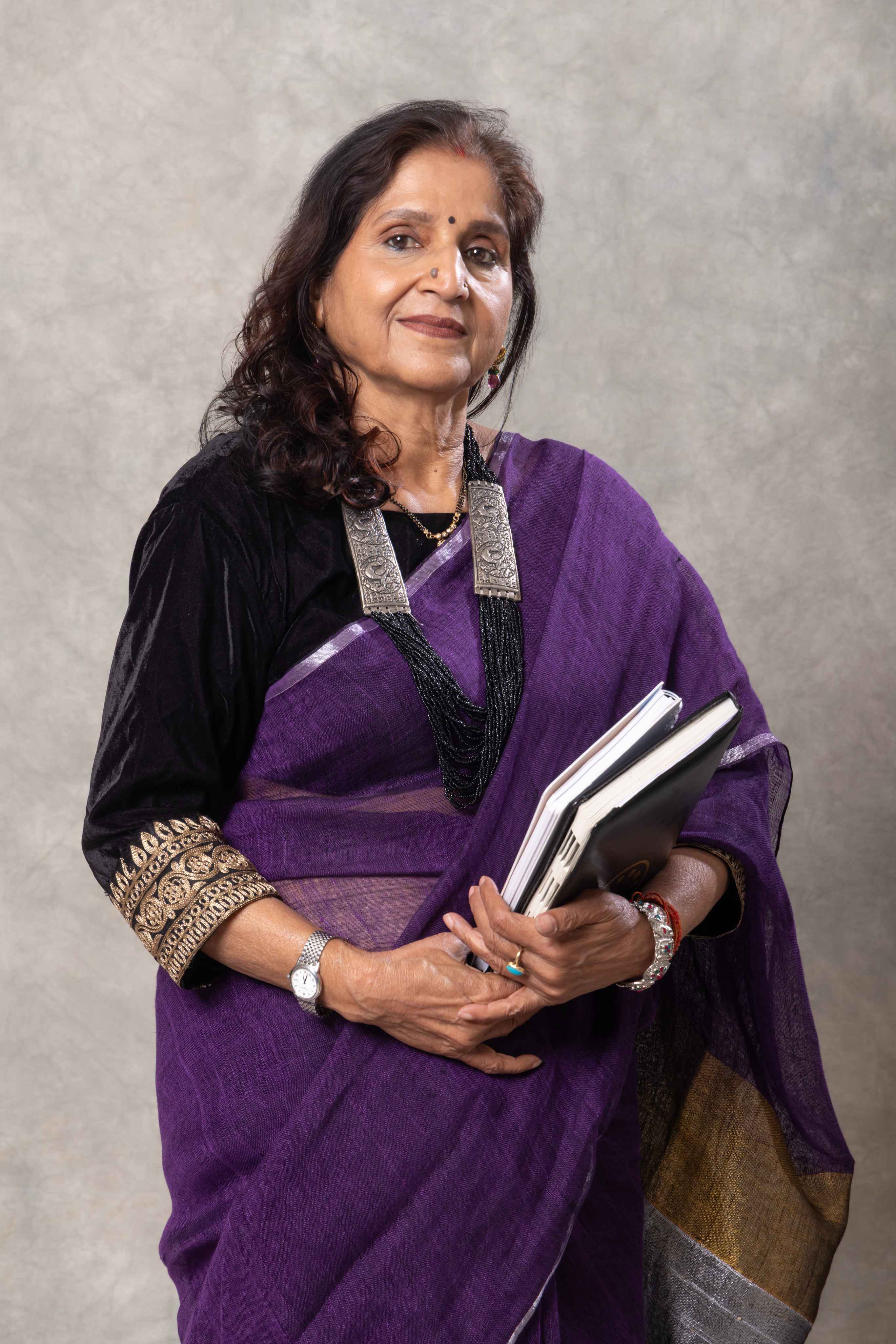 Woman in a purple saree holding books against a gray background