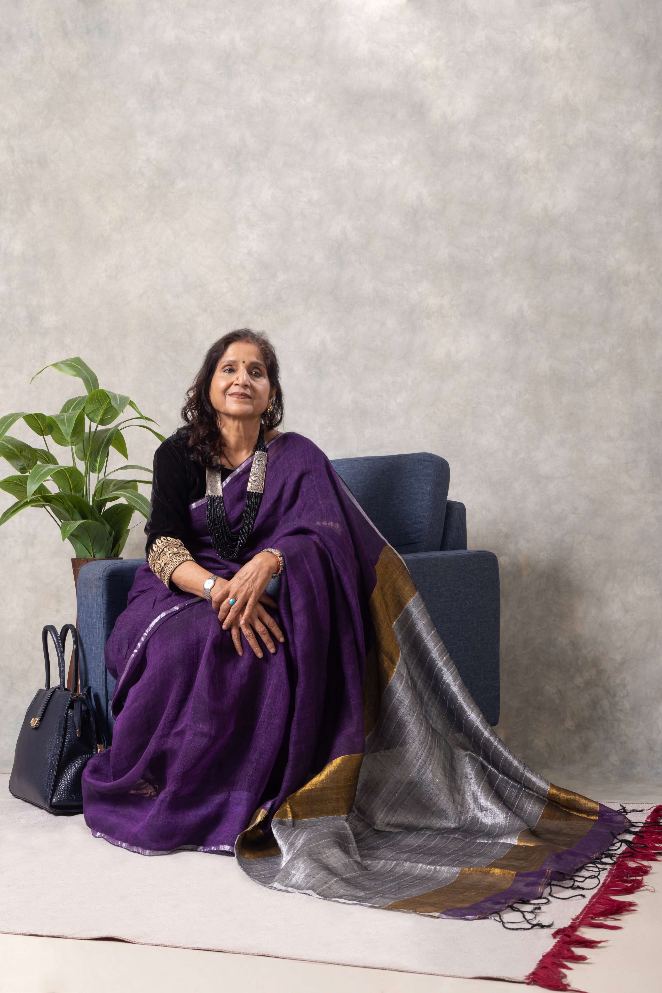 Woman in a purple saree sitting on a chair with a plant and handbag in the background