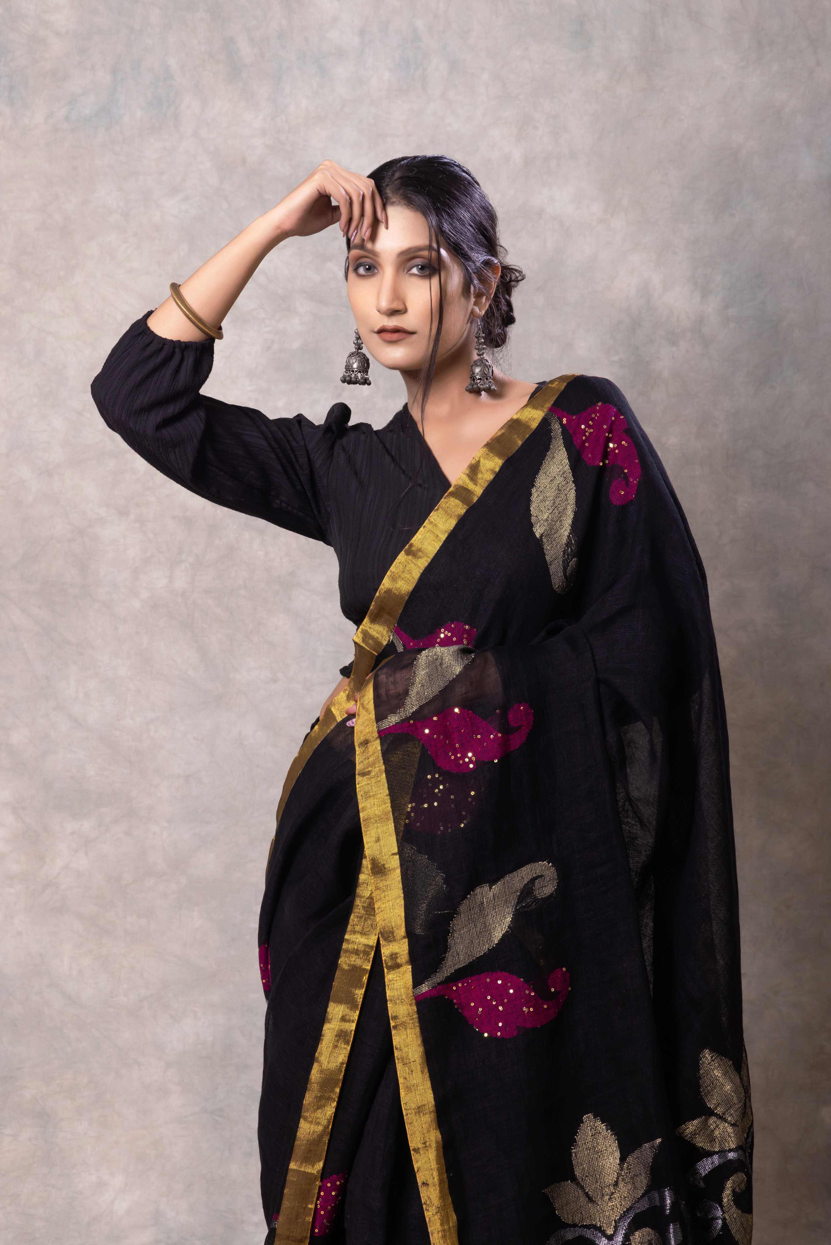 Woman wearing a black saree with gold and pink patterns against a gray background