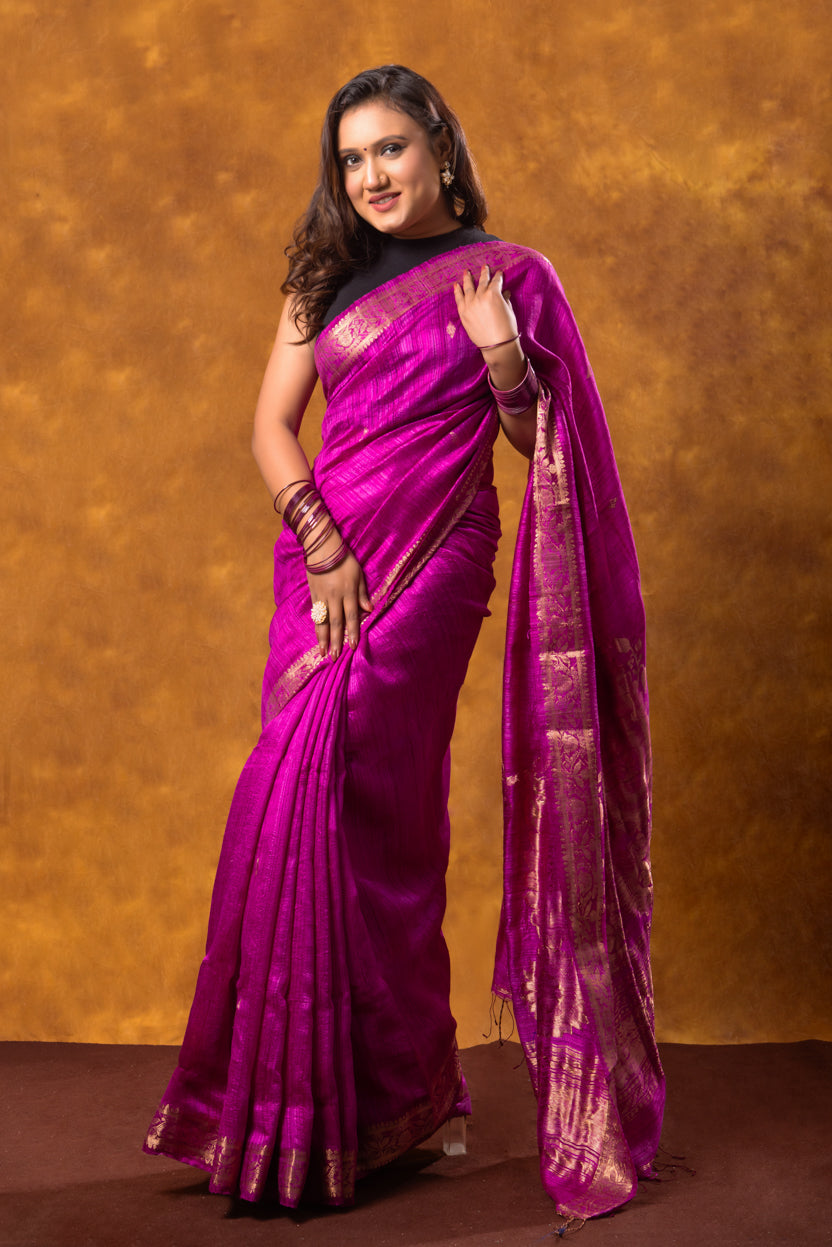 Woman wearing a vibrant purple saree with gold patterns against a warm brown background