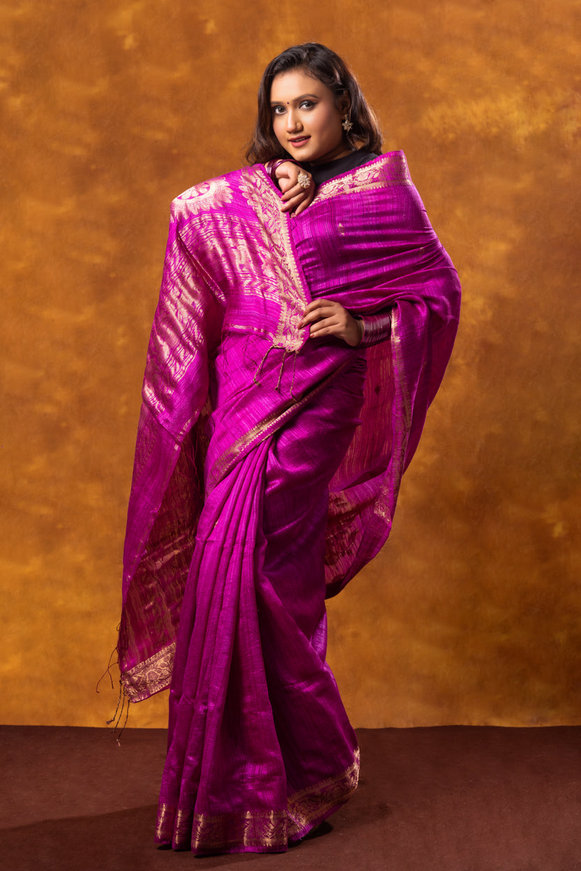 Woman wearing a vibrant purple saree with gold patterns against a brown background